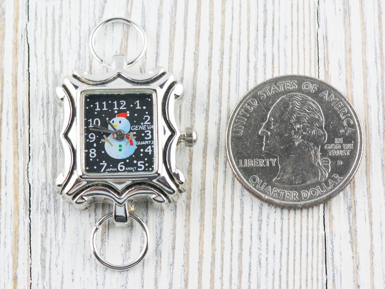 May include: A silver-toned pocket watch with a black face and a snowman design. The watch is about the size of a US quarter.