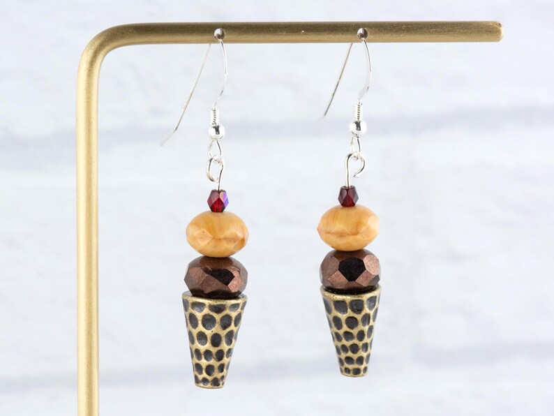 Puede incluir: Pendientes colgantes con dise&ntilde;o de cono de helado. Cada pendiente tiene un gancho plateado, una cuenta roja, una cuenta beige, una cuenta marr&oacute;n facetada y un cono dorado con un patr&oacute;n de puntos. Los pendientes se exhiben en un soporte dorado.