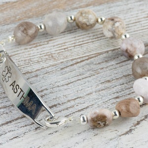May include: A silver medical alert bracelet with a silver tag that reads "ASTHMA" and a medical symbol. The bracelet is made with brown and white agate beads.