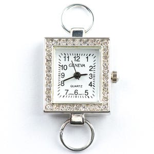 May include: A silver-toned Geneva quartz watch with a square face, surrounded by a border of small, clear crystals. The watch face is white with black numerals and hands. It has a ring at the top and bottom for attachment.
