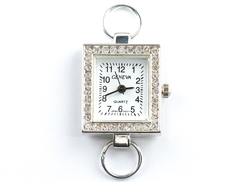 May include: A silver-toned Geneva quartz watch with a square face, surrounded by a border of small, clear crystals. The watch face is white with black numerals and hands. It has a ring at the top and bottom for attachment.
