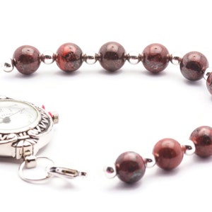 May include: A silver watch with a white face and black numbers is attached to a bracelet made of red and brown beads.