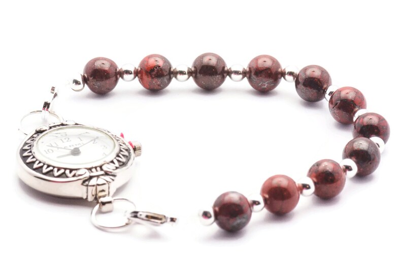 May include: A silver watch with a white face and black numbers is attached to a bracelet made of red and brown beads.