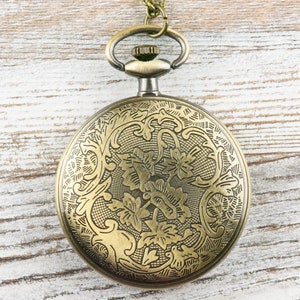 Op de afbeelding: Antieke messing zakhorloge met een sierlijk bloemmotief op de voorkant. Het horloge heeft een lus voor een ketting en een gedetailleerd patroon op de wijzerplaat. Het ligt op een lichte houten achtergrond.