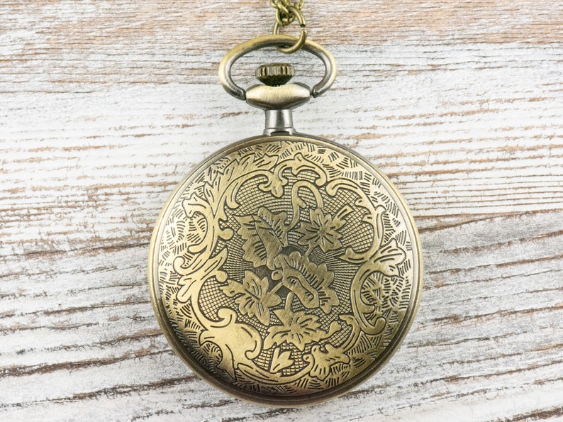 Op de afbeelding: Antieke messing zakhorloge met een sierlijk bloemmotief op de voorkant. Het horloge heeft een lus voor een ketting en een gedetailleerd patroon op de wijzerplaat. Het ligt op een lichte houten achtergrond.