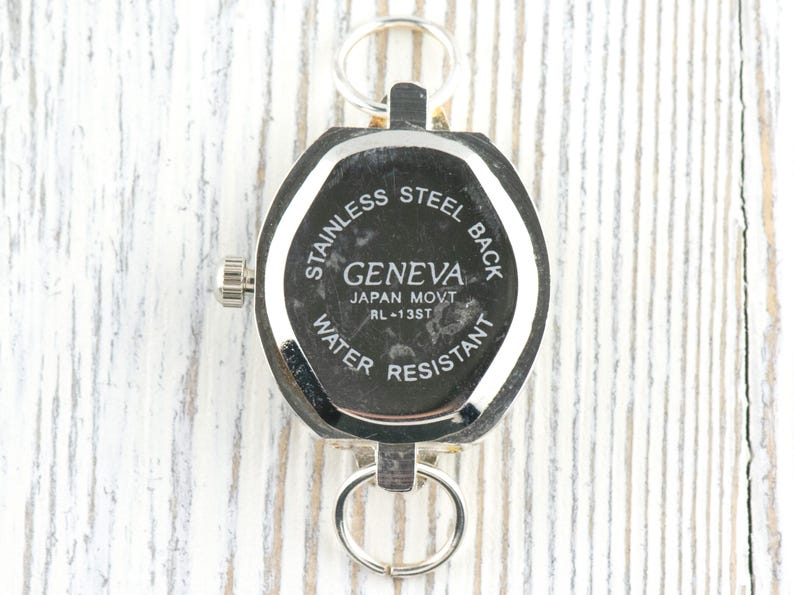May include: A silver-toned Geneva watch with a stainless steel back and water-resistant design. The watch has a hexagonal shape and two key rings. The text "GENEVA JAPAN MOVT RL-13ST" is visible.