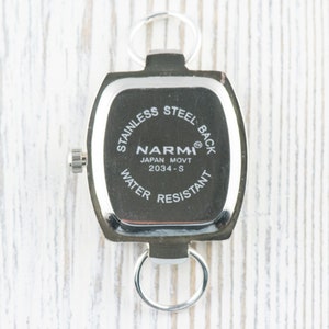 May include: The back of a silver watch with a square face. The watch face has the text "STAINLESS STEEL BACK", "NARM&reg;", "JAPAN MOVT 2034-S", and "WATER RESISTANT".