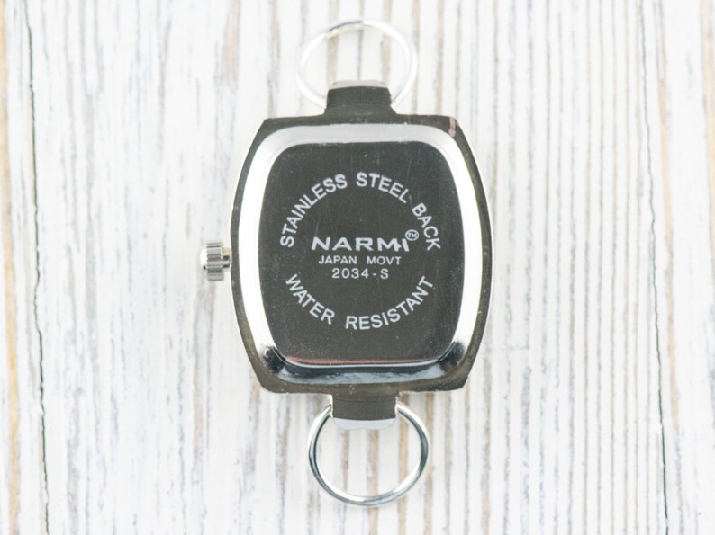 May include: The back of a silver watch with a square face. The watch face has the text "STAINLESS STEEL BACK", "NARM&reg;", "JAPAN MOVT 2034-S", and "WATER RESISTANT".