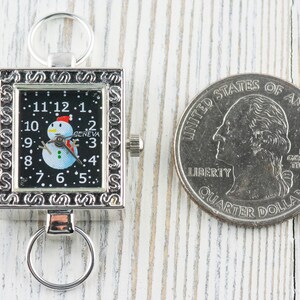 K&ouml;nnte beinhalten: Eine silberne Taschenuhr mit schwarzem Zifferblatt und einem Schneemann-Motiv. Die Uhr hat eine silberne Kette und ein quadratisches Zifferblatt mit einem dekorativen Rand. Die Uhr liegt neben einer US-amerikanischen 25-Cent-M&uuml;nze.