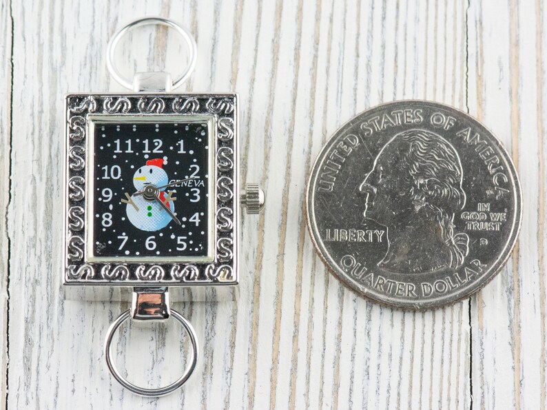 K&ouml;nnte beinhalten: Eine silberne Taschenuhr mit schwarzem Zifferblatt und einem Schneemann-Motiv. Die Uhr hat eine silberne Kette und ein quadratisches Zifferblatt mit einem dekorativen Rand. Die Uhr liegt neben einer US-amerikanischen 25-Cent-M&uuml;nze.