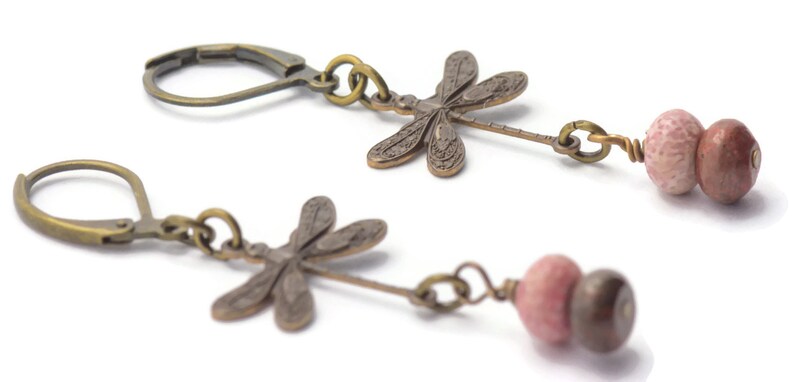 Puede incluir: Un par de pendientes colgantes con un dije de lib&eacute;lula y dos cuentas de color rosa y marr&oacute;n. Los dijes de lib&eacute;lula est&aacute;n hechos de metal y tienen un acabado bronce. Las cuentas est&aacute;n hechas de piedra y tienen un acabado natural.