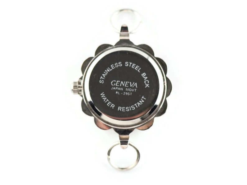 May include: A silver-toned Geneva watch with a floral-shaped bezel and two key rings. The watch back is black with white text that reads "STAINLESS STEEL BACK", "GENEVA JAPAN MOVT RL-255T", and "WATER RESISTANT".