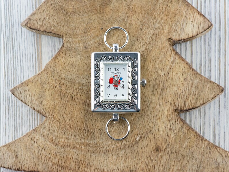 May include: A silver pocket watch with a white face and a Santa Claus design. The watch has a decorative border and is attached to a silver chain.