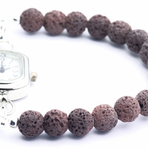 Puede incluir: Un reloj plateado con esfera blanca y n&uacute;meros negros est&aacute; unido a una pulsera hecha de cuentas de roca volc&aacute;nica marr&oacute;n oscuro y porosa. El reloj tiene una esfera rectangular con bordes redondeados. La pulsera es curva.