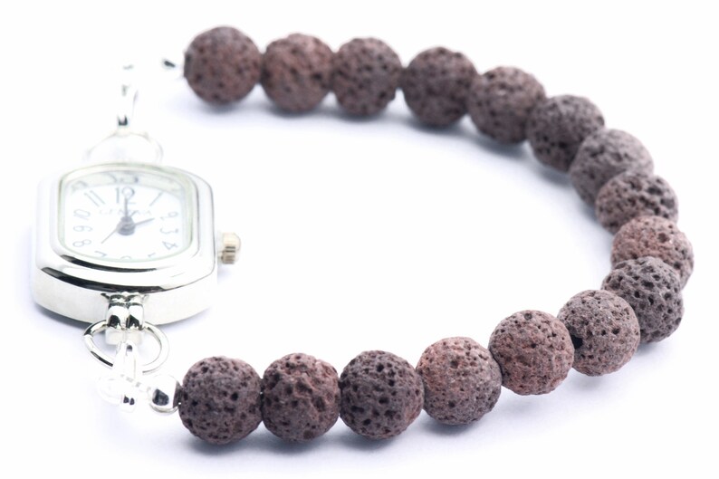 Puede incluir: Un reloj plateado con esfera blanca y n&uacute;meros negros est&aacute; unido a una pulsera hecha de cuentas de roca volc&aacute;nica marr&oacute;n oscuro y porosa. El reloj tiene una esfera rectangular con bordes redondeados. La pulsera es curva.