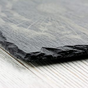 K&ouml;nnte beinhalten: Nahaufnahme einer schwarzen Schiefer-Servierplatte mit einer rauen, strukturierten Oberfl&auml;che. Die Platte ist angewinkelt und die Kanten sind leicht abgebrochen.