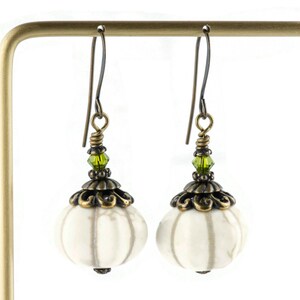 May include: White ceramic earrings with a textured surface, bronze filigree accents, and green crystal beads hanging from bronze hooks.
