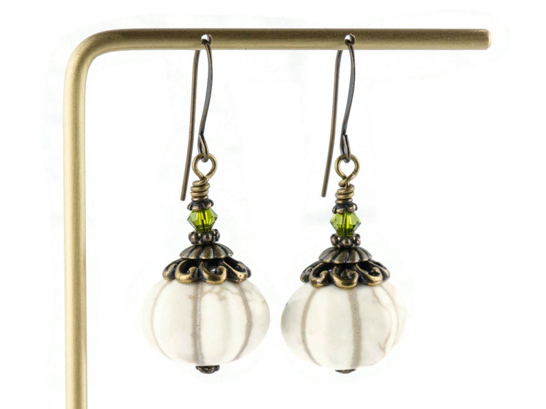 May include: White ceramic earrings with a textured surface, bronze filigree accents, and green crystal beads hanging from bronze hooks.