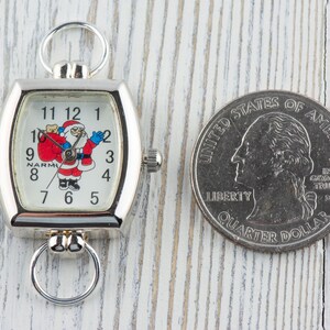 May include: A silver pocket watch with a square face featuring a Santa Claus illustration and the numbers 1 through 12. The watch is about the size of a US quarter.