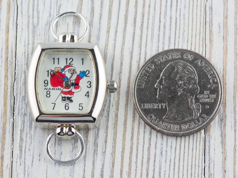 May include: A silver pocket watch with a square face featuring a Santa Claus illustration and the numbers 1 through 12. The watch is about the size of a US quarter.