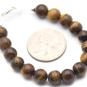 Op de afbeelding: Een bruine en gouden tijgeroog kralen armband met een zilveren sluiting. De armband ligt naast een Amerikaanse kwart voor referentie van de grootte.