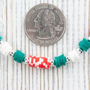 K&ouml;nnte beinhalten: Nahaufnahme eines Armbands aus wei&szlig;en, gr&uuml;nen und roten Perlen. Das Armband liegt auf einer wei&szlig;en Holzoberfl&auml;che mit einer US-amerikanischen 25-Cent-M&uuml;nze als Gr&ouml;&szlig;envergleich.