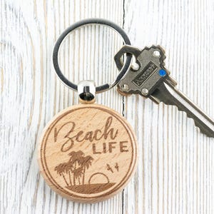 Pu&ograve; includere: Un portachiavi in legno con il testo "Beach Life" inciso su di esso. Il portachiavi presenta un design di palme e un tramonto. Il portachiavi &egrave; attaccato a un portachiavi d'argento.