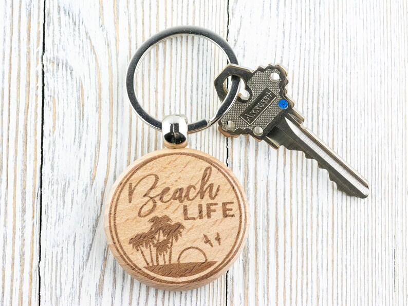 Pu&ograve; includere: Un portachiavi in legno con il testo "Beach Life" inciso su di esso. Il portachiavi presenta un design di palme e un tramonto. Il portachiavi &egrave; attaccato a un portachiavi d'argento.