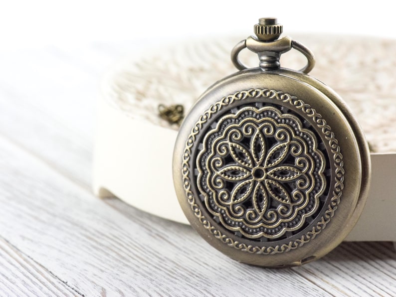 K&ouml;nnte beinhalten: Antike bronzefarbene Taschenuhr mit einem verzierten Blumendesign auf der Vorderseite. Die Uhr hat eine runde Form mit einem detaillierten, durchbrochenen Muster. Sie befindet sich vor einem wei&szlig;en Hintergrund.