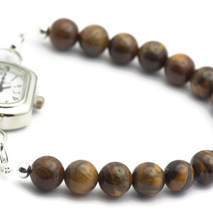 Op de afbeelding: Een zilveren horloge met een vierkante wijzerplaat en bruine tijgeroog kralen bevestigd aan de band.