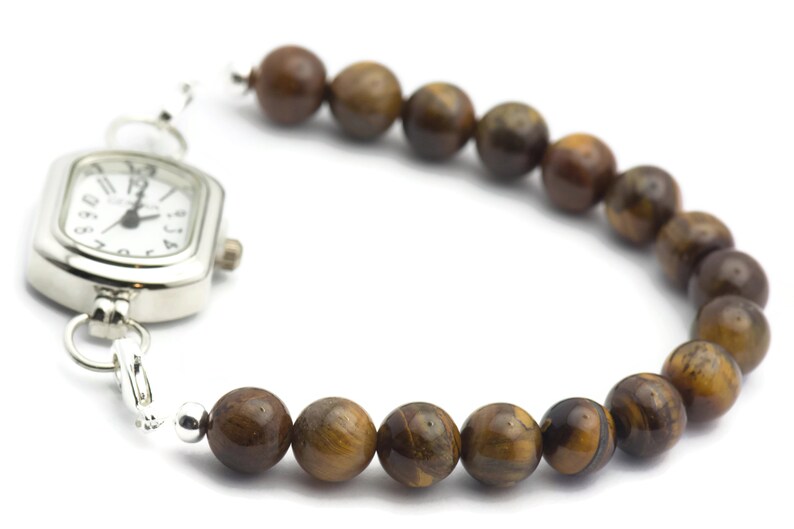 Op de afbeelding: Een zilveren horloge met een vierkante wijzerplaat en bruine tijgeroog kralen bevestigd aan de band.