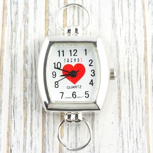 Puede incluir: Reloj de cuarzo plateado con esfera blanca, n&uacute;meros negros y un coraz&oacute;n rojo. El reloj tiene forma rectangular con bordes redondeados y dos lazos circulares. La palabra "TRENDZ" est&aacute; impresa encima del coraz&oacute;n.