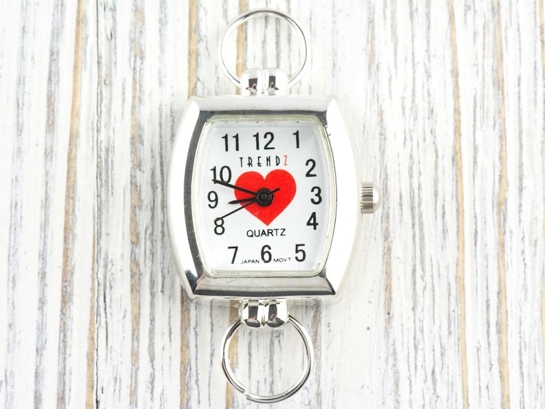 Puede incluir: Reloj de cuarzo plateado con esfera blanca, n&uacute;meros negros y un coraz&oacute;n rojo. El reloj tiene forma rectangular con bordes redondeados y dos lazos circulares. La palabra "TRENDZ" est&aacute; impresa encima del coraz&oacute;n.