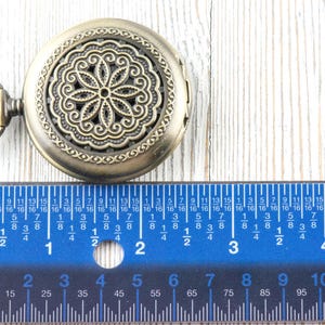 K&ouml;nnte beinhalten: Antike bronzefarbene Taschenuhr mit einem verzierten Blumendesign auf dem Deckel. Ein blaues Lineal mit Zentimetermarkierungen befindet sich darunter. Die Uhr hat einen Durchmesser von etwa 5 cm.