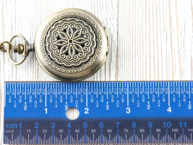 K&ouml;nnte beinhalten: Antike bronzefarbene Taschenuhr mit einem verzierten Blumendesign auf dem Deckel. Ein blaues Lineal mit Zentimetermarkierungen befindet sich darunter. Die Uhr hat einen Durchmesser von etwa 5 cm.
