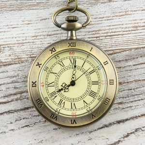 Op de afbeelding: Antieke messing zakhorloge met een cr&egrave;mekleurige wijzerplaat en zwarte Romeinse cijfers. Het horloge heeft een gedetailleerd ontwerp en een ketting. De wijzers staan ongeveer op 12:00 uur. Het horloge ligt op een witte houten achtergrond.