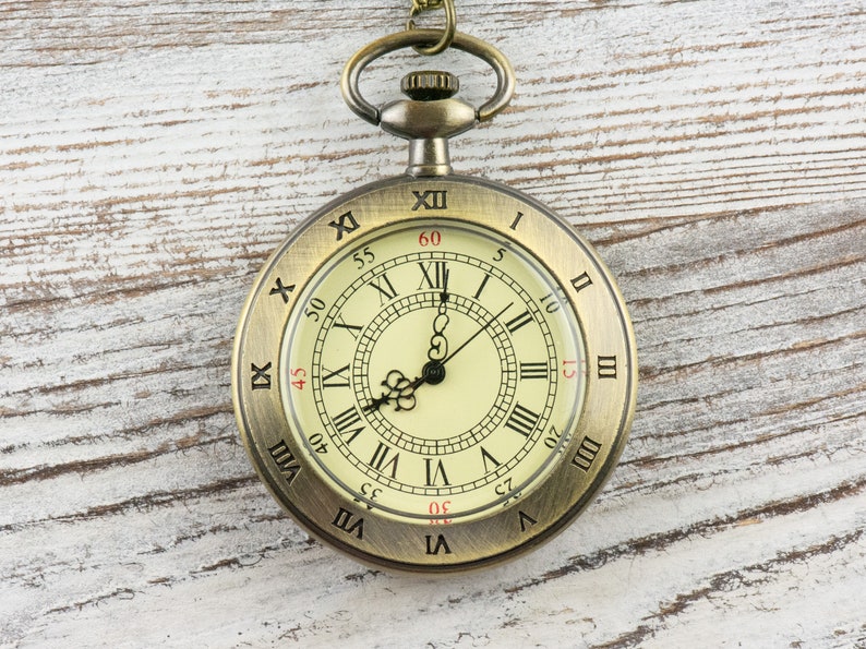Op de afbeelding: Antieke messing zakhorloge met een cr&egrave;mekleurige wijzerplaat en zwarte Romeinse cijfers. Het horloge heeft een gedetailleerd ontwerp en een ketting. De wijzers staan ongeveer op 12:00 uur. Het horloge ligt op een witte houten achtergrond.