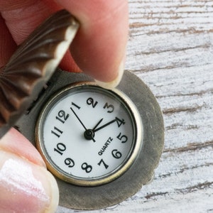 May include: A small, antique-looking pocket watch with a round, white face and black Roman numeral markings. The watch has a brown metal casing with a decorative, scalloped lid.