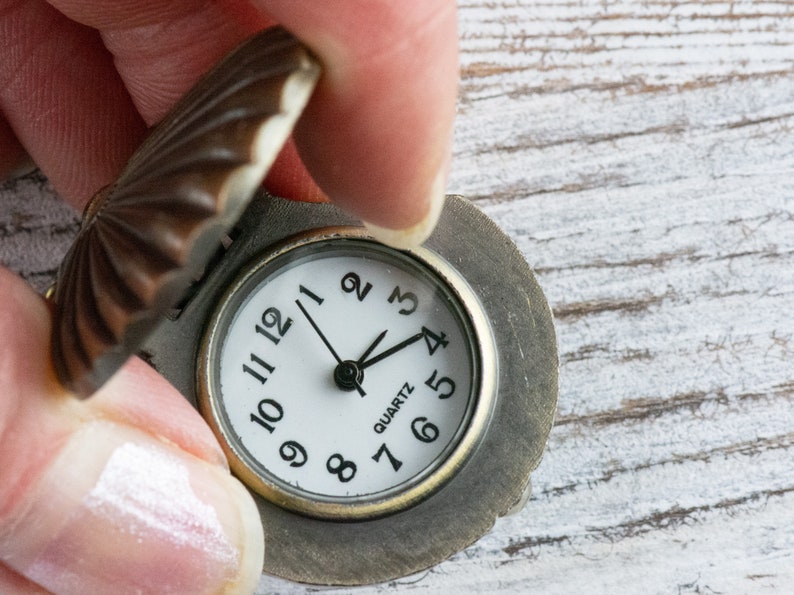 May include: A small, antique-looking pocket watch with a round, white face and black Roman numeral markings. The watch has a brown metal casing with a decorative, scalloped lid.