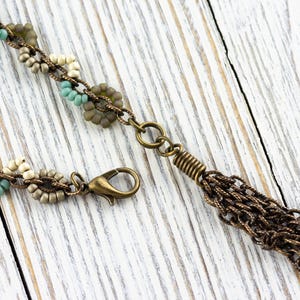 May include: A beaded necklace with a bronze chain tassel. The necklace features small, round beads in shades of beige, brown, and turquoise.