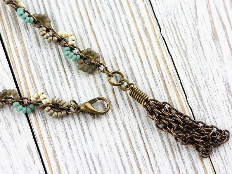May include: A beaded necklace with a bronze chain tassel. The necklace features small, round beads in shades of beige, brown, and turquoise.