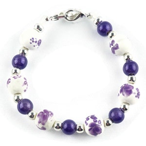 May include: A purple and white beaded bracelet with a silver clasp. The beads are round and have a floral design.