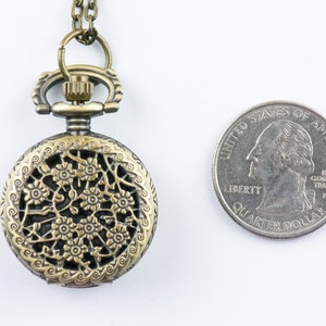 K&ouml;nnte beinhalten: Antiker, bronzefarbener Taschenuhr-Anh&auml;nger mit floralem Filigranmuster. Die Uhr ist rund und hat eine Kette. Eine amerikanische Vierteldollarm&uuml;nze liegt neben der Uhr zum Gr&ouml;&szlig;envergleich.