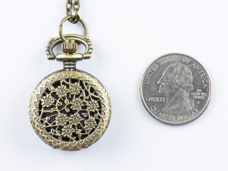 K&ouml;nnte beinhalten: Antiker, bronzefarbener Taschenuhr-Anh&auml;nger mit floralem Filigranmuster. Die Uhr ist rund und hat eine Kette. Eine amerikanische Vierteldollarm&uuml;nze liegt neben der Uhr zum Gr&ouml;&szlig;envergleich.