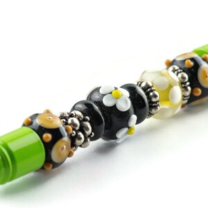 May include: A green pen with black, white, and yellow glass beads. The beads have a daisy pattern.