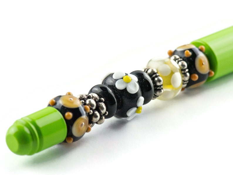 May include: A green pen with black, white, and yellow glass beads. The beads have a daisy pattern.
