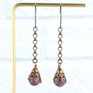 May include: A pair of drop earrings with a vintage style. Each earring has a small, round, purple gemstone in a filigree teardrop setting. The earrings hang from a chain of heart-shaped links.