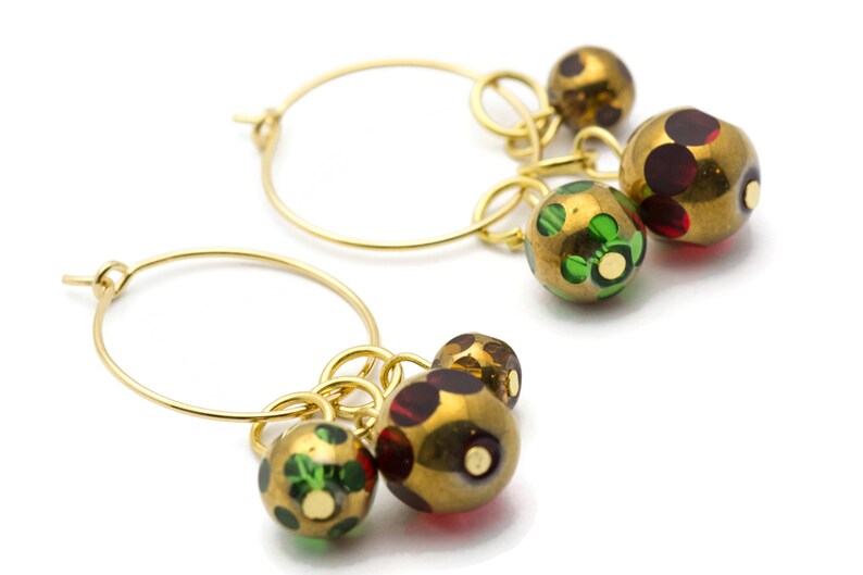 May include: Gold hoop earrings with dangling charms. The charms are round and have a gold base with green and red accents.