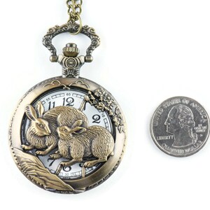 Puede incluir: Reloj de bolsillo antiguo de bronce con un dise&ntilde;o detallado de conejos. La esfera del reloj es blanca con n&uacute;meros y manecillas negras. Una moneda de veinticinco centavos se encuentra junto al reloj para comparar el tama&ntilde;o.