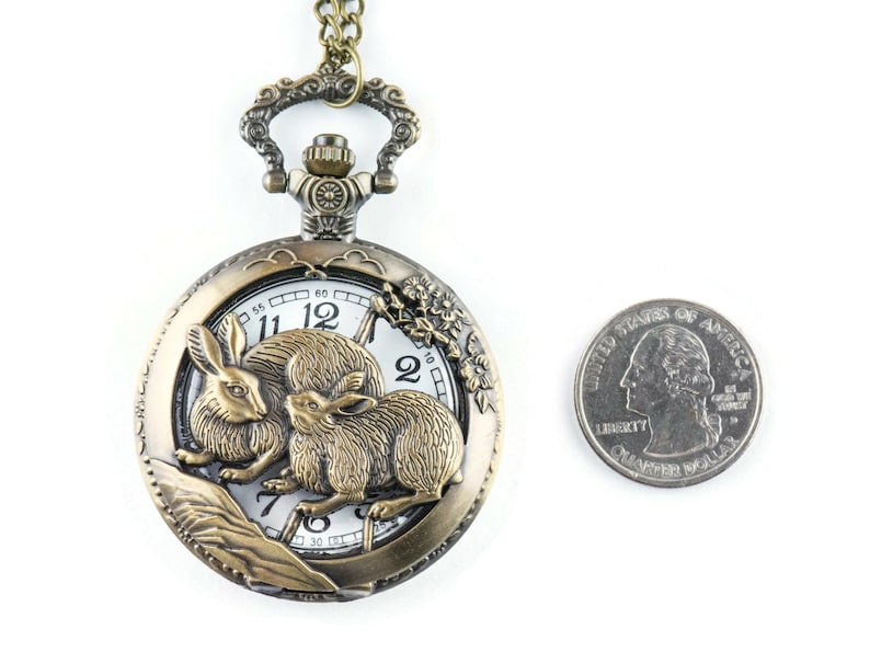 Puede incluir: Reloj de bolsillo antiguo de bronce con un dise&ntilde;o detallado de conejos. La esfera del reloj es blanca con n&uacute;meros y manecillas negras. Una moneda de veinticinco centavos se encuentra junto al reloj para comparar el tama&ntilde;o.